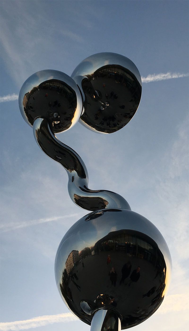 Three polished stainless steel spheres with curved connecting element against sky with condensation trails